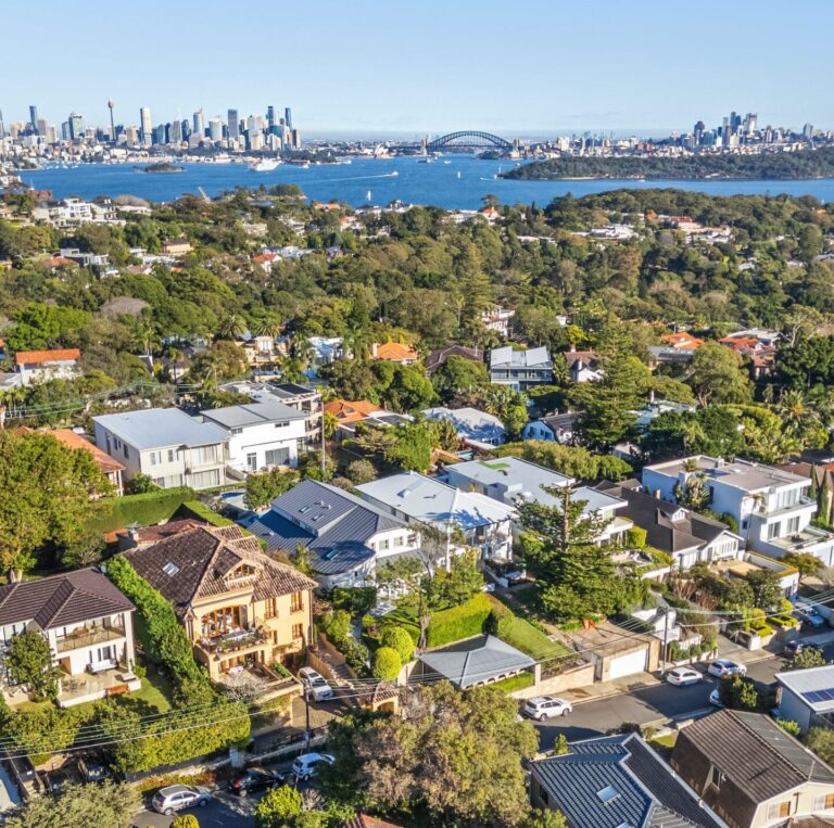 Aerial view of a leafy suburban neighborhood with houses, trees, and cars; beyond the suburb is a blue harbor, with a city skyline and an arched bridge visible in the background under a clear sky.