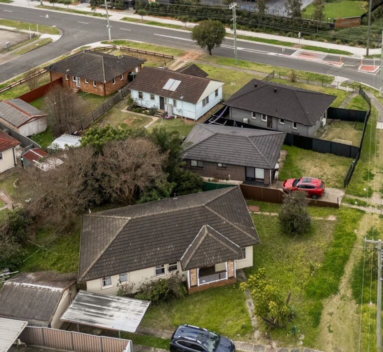 Aerial view of a suburban neighborhood with several single-story houses, green lawns, a red car parked in a driveway, and intersecting streets surrounding the properties.