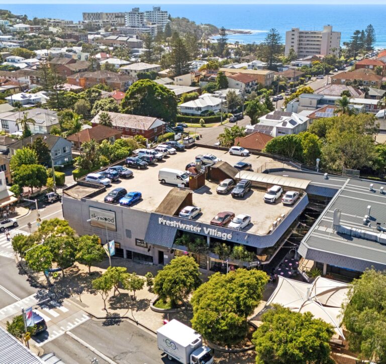 Aerial view of Freshwater Village shopping center with cars parked on its rooftop, surrounded by trees and residential buildings, with the coastline and ocean visible in the background.