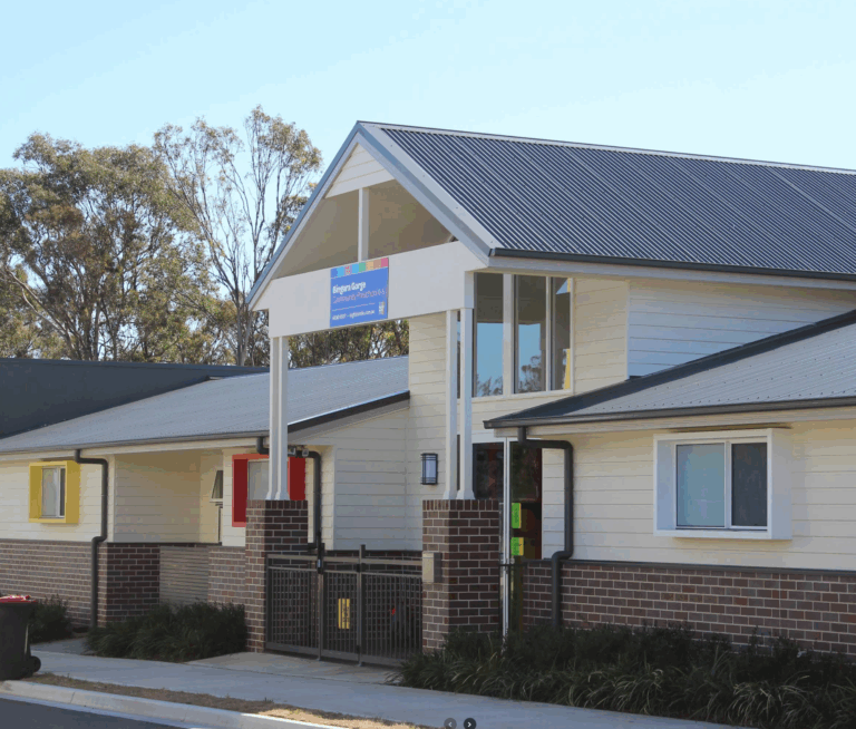 Single-story modern building with cream siding, brick foundation, and dark roof. A sign above the entrance suggests its a community or healthcare center. Trees are visible in the background under a clear sky.