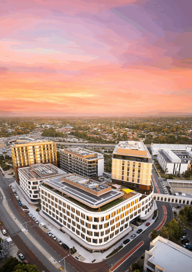 Aerial view of a modern multi-story building complex at sunset, with roads and parked cars surrounding it, and a sprawling suburban area extending into the distance under a colorful sky.