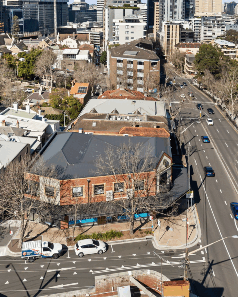 Aerial view of a city intersection with cars parked and driving on the streets, surrounded by multi-story buildings, trees, and a mix of modern and older architecture.