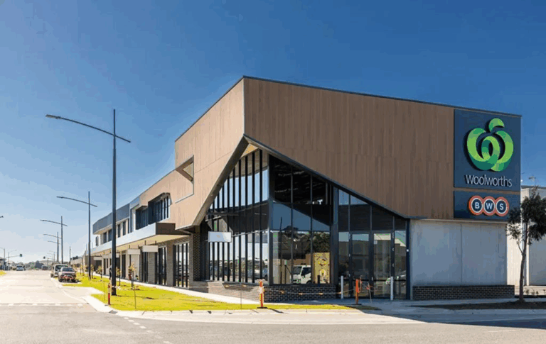 A modern Woolworths supermarket building with large glass windows and a distinctive angular roof, located on a street corner under a clear blue sky. The BWS logo is also displayed on the building.