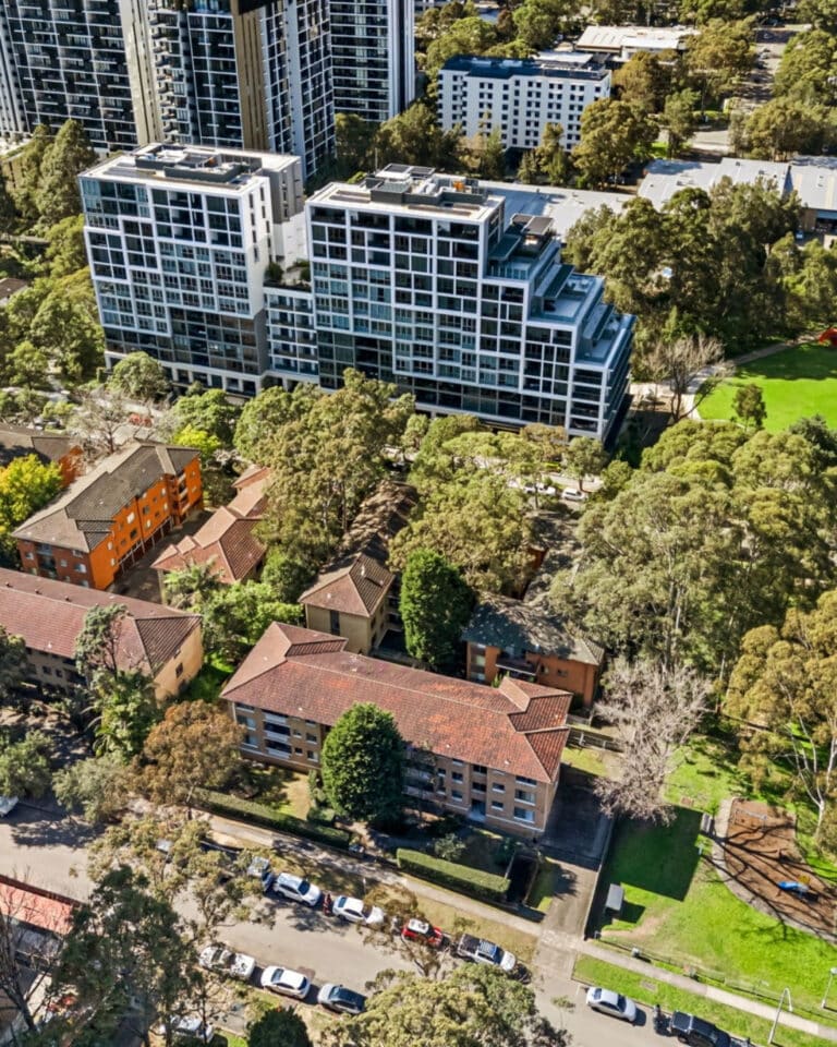 Aerial view of a city neighborhood with modern high-rise apartment buildings, older low-rise brick apartments, tree-lined streets, parked cars, and green spaces with dense trees.