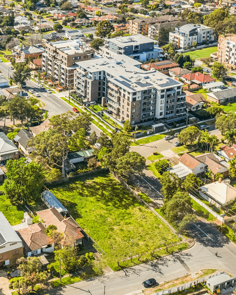 Aerial view of a suburban neighborhood with modern apartment buildings, single-family homes, green lawns, and tree-lined streets on a sunny day.